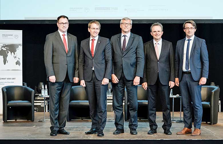 Fünf Herren in grauen Anzügen stehen nebeneinander auf einer Bühne und schauen in Richtung der Kamera.